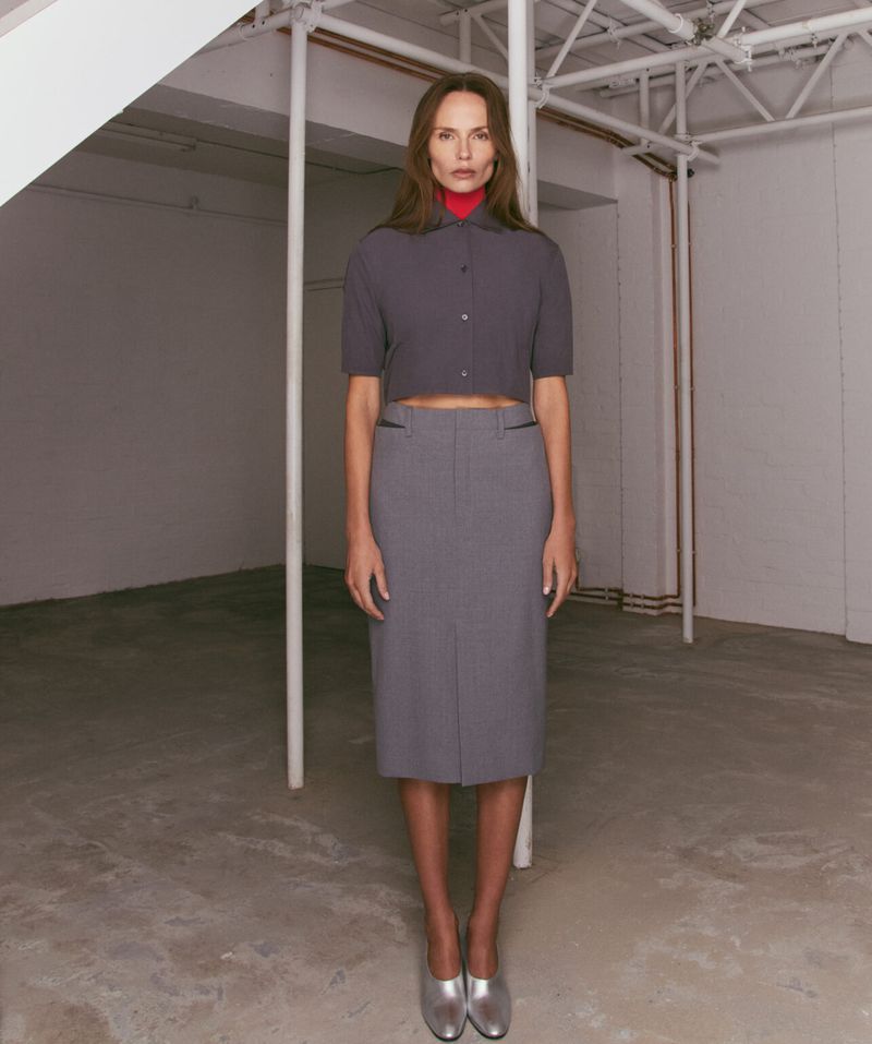 Full-length shot of a stylish woman posing in a gray skirt and top, complemented by metallic silver shoes in an indoor location.