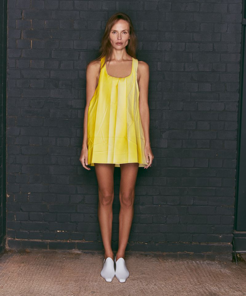 Full shot of an adult woman standing in a bright yellow dress and white shoes, in front of a dark brick wall. Fashion shoot.