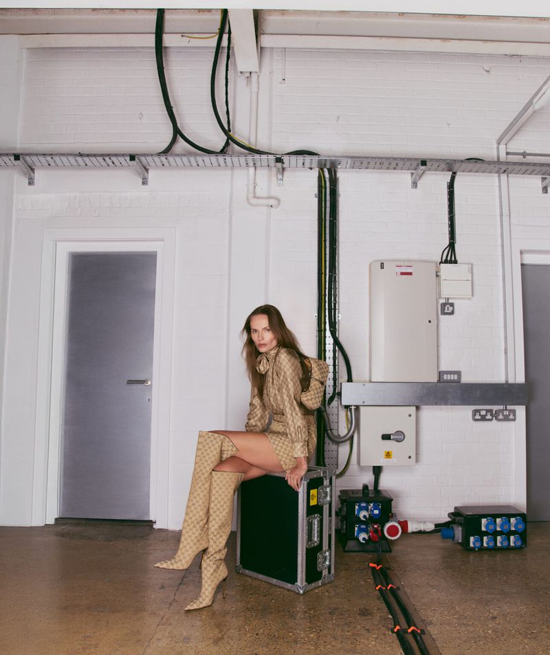 A woman wearing patterned thigh-high boots and matching jacket sits on a black road case in an industrial-style white room with exposed wiring.