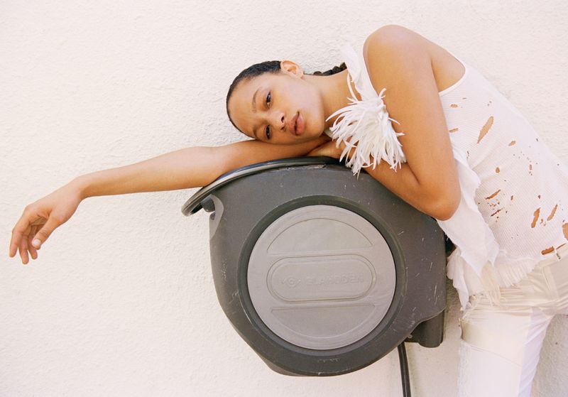 A woman in a white top and pants leans against a grey Gardena hose reel against a white wall, with a relaxed pose.