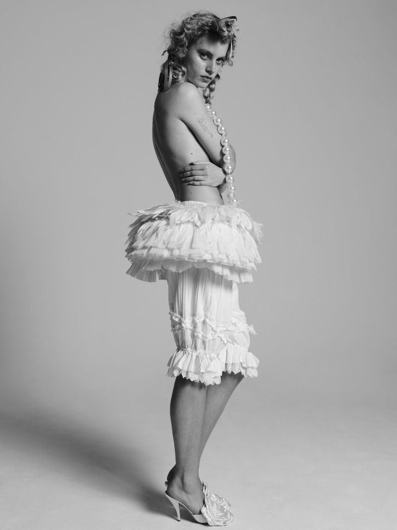 A black and white portrait of a woman with curly hair, pearls, and a frilly white costume posing in a photo studio.