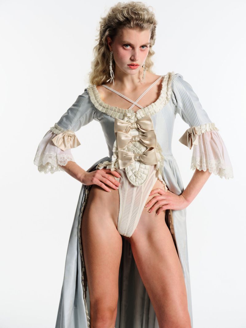 Blonde woman in a blue 18th-century-style dress with a large bow poses against a white background in a studio shot, looking at the camera.