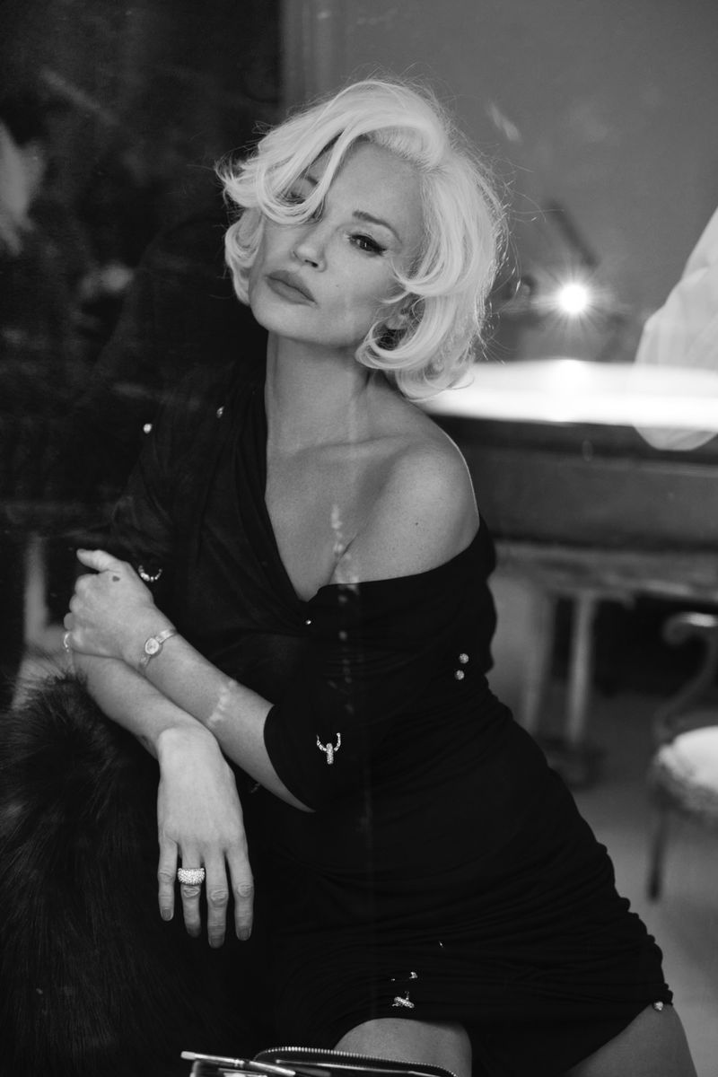 Monochrome portrait of a blonde adult female seated in a black dress with jewelry. Shallow depth of field, indoor environment.