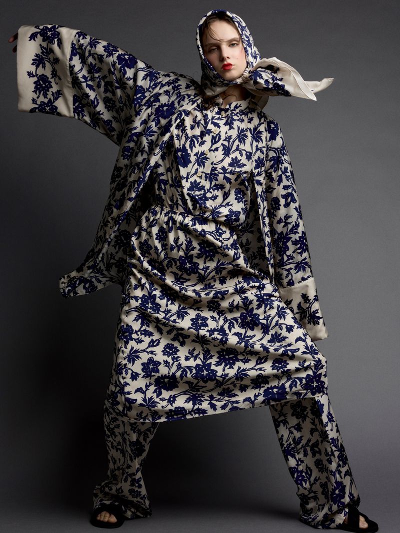 A woman wearing a cream colored pantsuit with a blue floral pattern and a matching headscarf poses in a photo studio against gray backdrop.