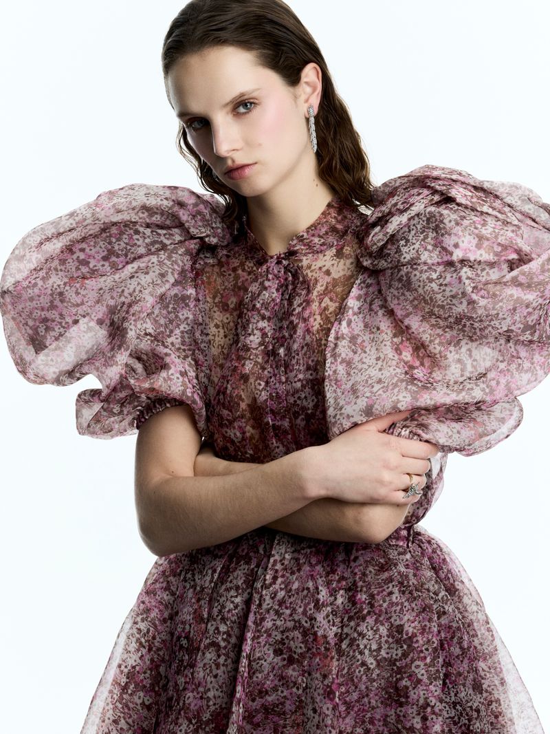 A woman with fair skin wears a semi-sheer purple floral dress with full, puffed sleeves against a clean, bright white background.