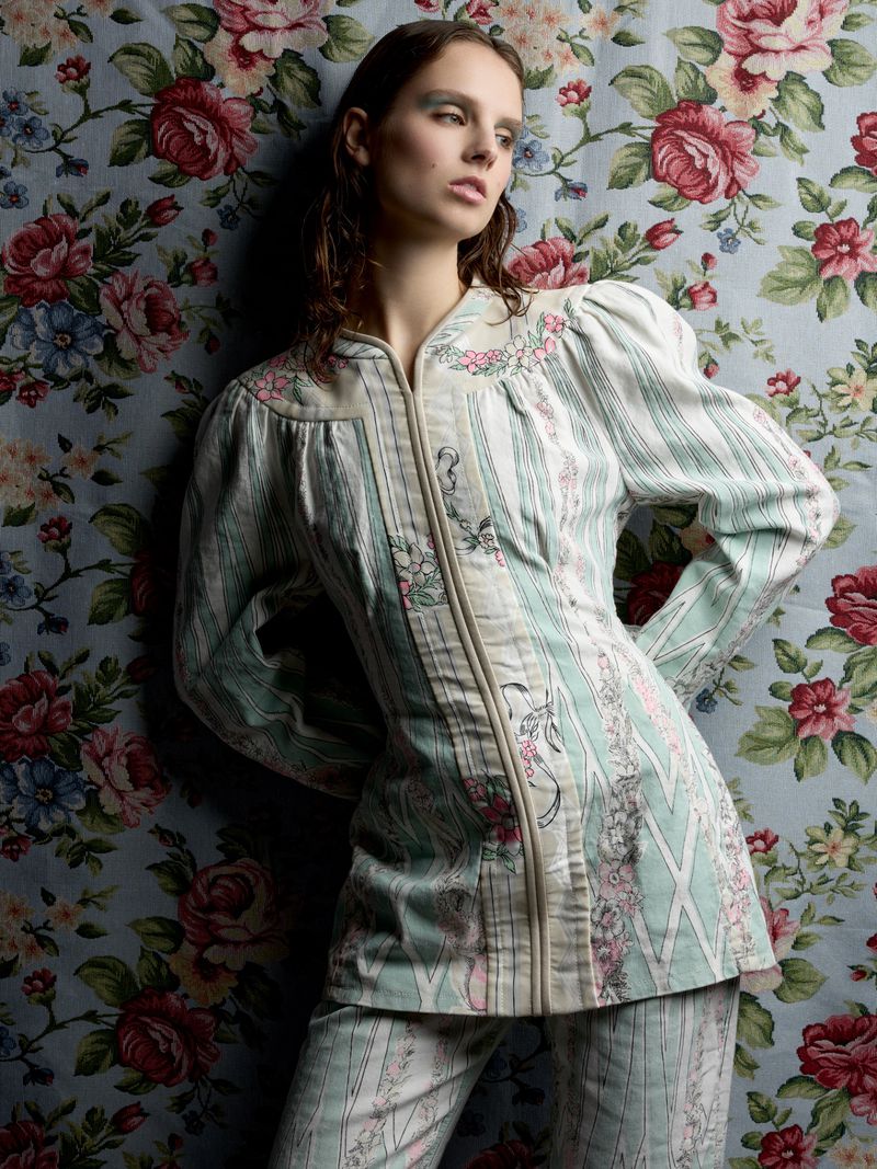 A model poses in a light blue floral patterned jacket and pants, with a matching floral background, in a fashion studio shot.