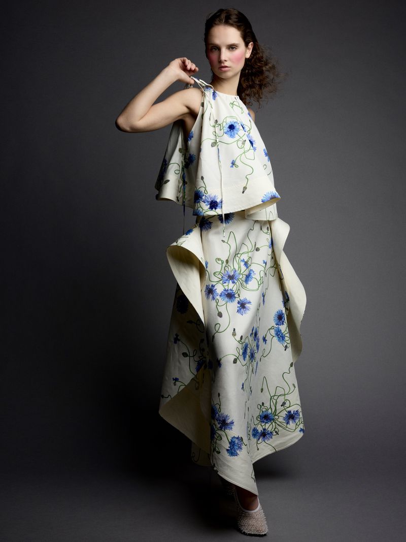 Woman in a flowing, two-piece, ivory-colored dress with blue floral patterns posing against a dark gray background in a studio setting.