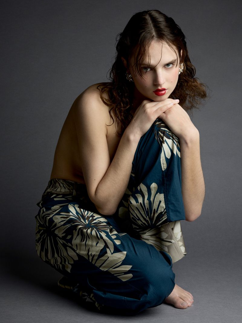 A female model in floral pants poses in a studio with a grey background. She is crouched down, facing forward with bright red lipstick.