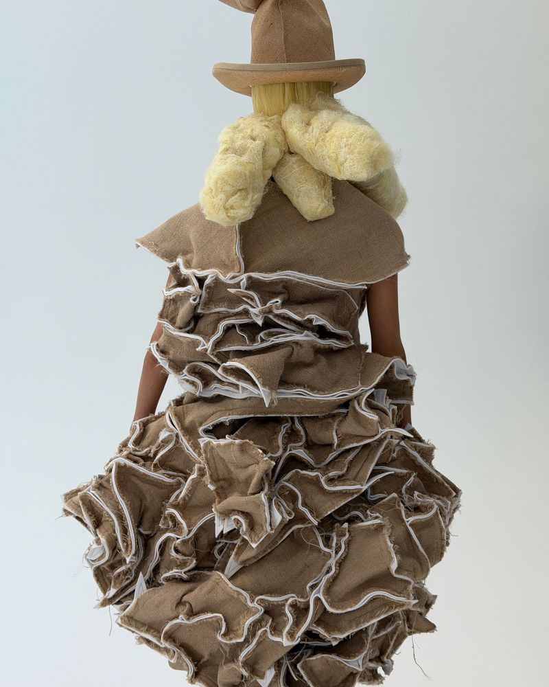 Rear view of a person wearing a whimsical burlap dress with ruffles, a pointed hat, and textured blonde hair in a white studio.