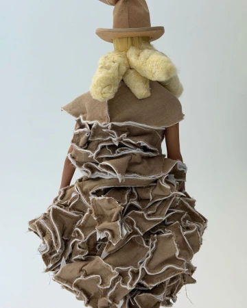 Rear view of a person wearing a whimsical burlap dress with ruffles, a pointed hat, and textured blonde hair in a white studio.