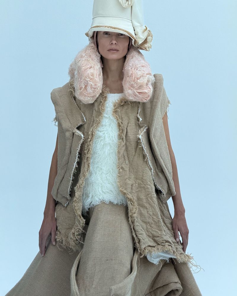 Woman in avant-garde fashion featuring a sleeveless burlap vest, long skirt, and sculpted beige hat against a light background.