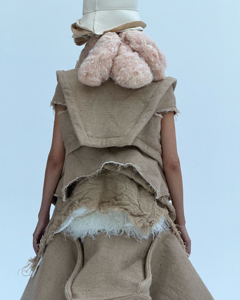 A person from the back wearing a deconstructed tan fabric dress, a beige bucket hat, and a pink furry object over the shoulders.