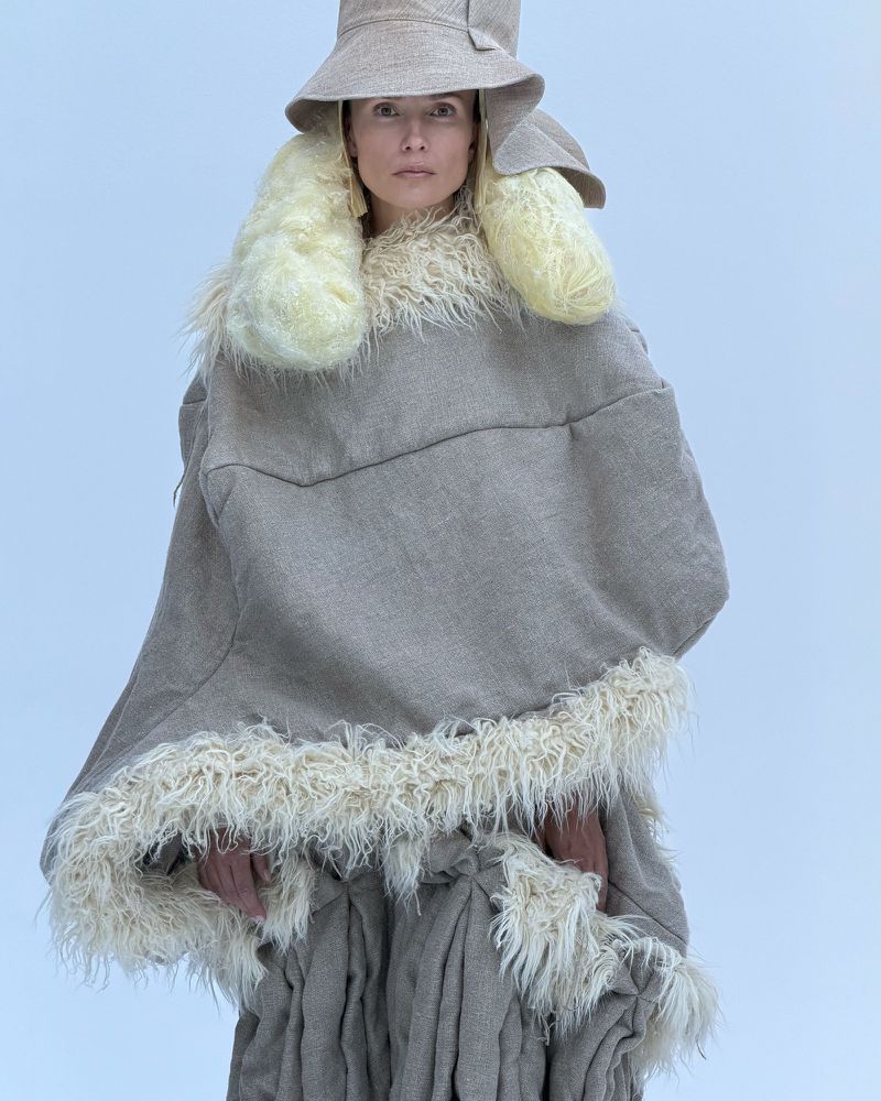 A woman poses in a neutral-tone hat and poncho with fur trim against a light background, showcasing unique fashion and textures.
