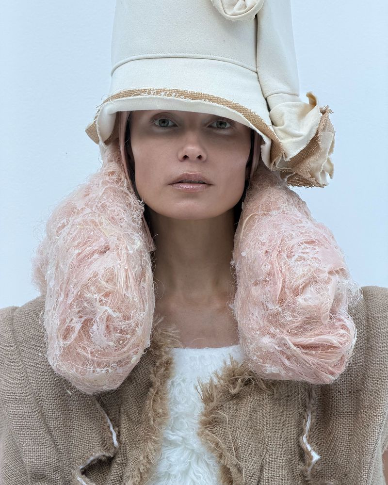 Avant-garde fashion portrait of a woman wearing a beige hat, oversized textured earrings, and burlap top against a white backdrop.