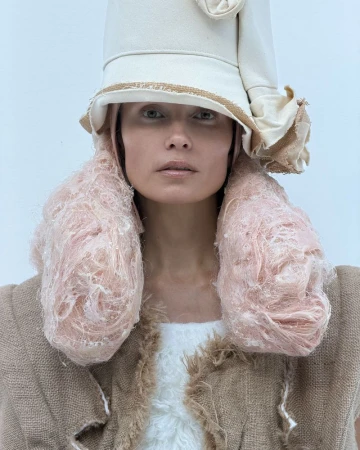 Avant-garde fashion portrait of a woman wearing a beige hat, oversized textured earrings, and burlap top against a white backdrop.