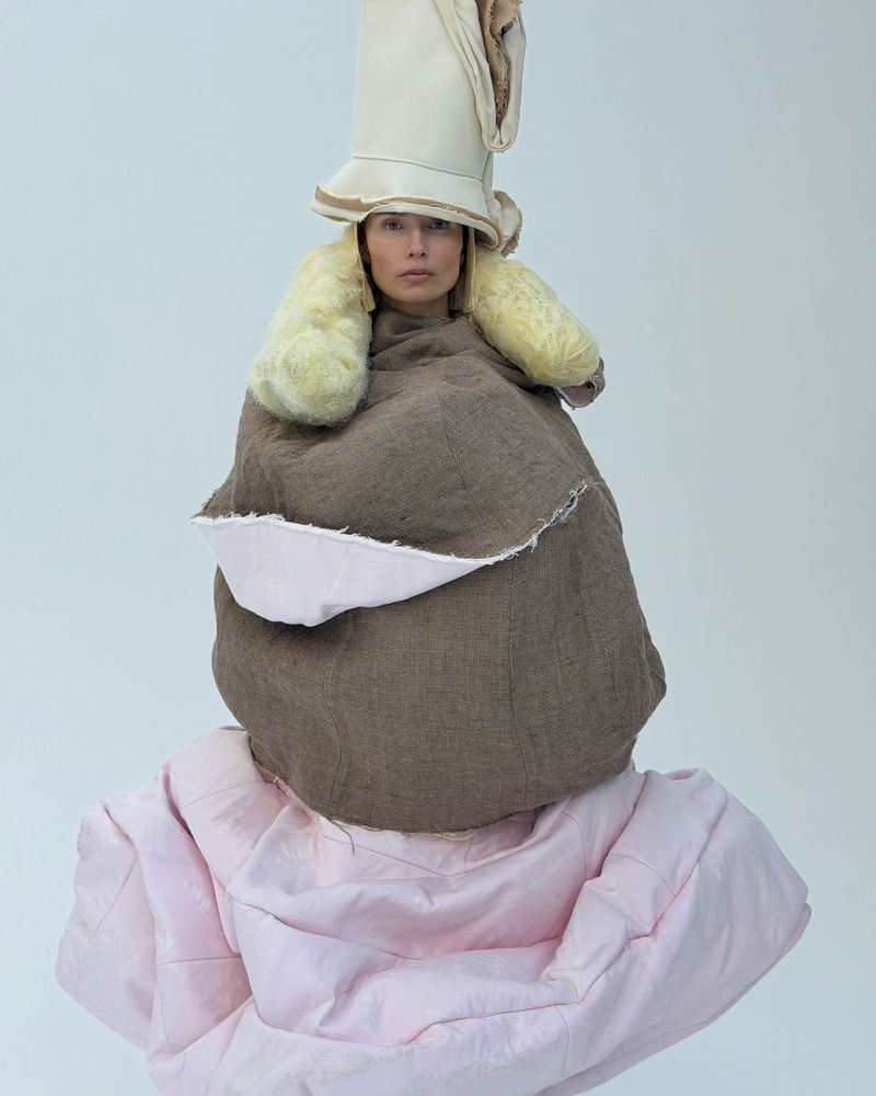 Adult female in avant-garde fashion outfit with beige hat, brown shawl, and pink skirt posing in a neutral studio against a white backdrop.