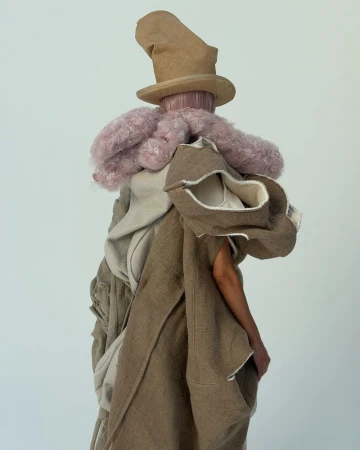 Person in conceptual fashion ensemble featuring a light-brown hat, pink faux-fur collar, and multi-layered beige and brown draped garment.