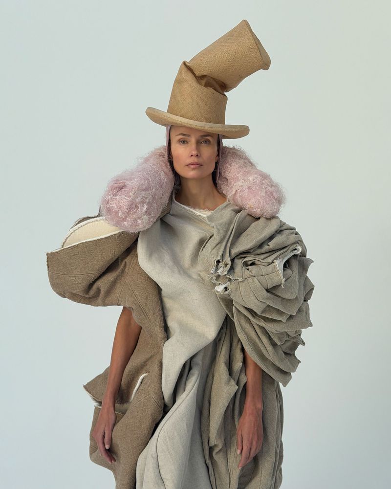 Elegant woman wearing a sculptural hat and draped fabric in a studio setting, showcasing unique fashion and textile art against a white background.