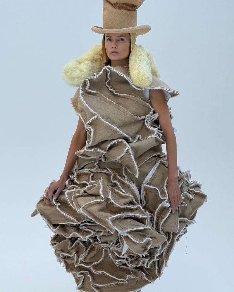 A woman models avant-garde fashion on a white backdrop, featuring an elaborate ruffled dress and matching hat made of rough burlap.