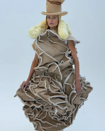 A woman models avant-garde fashion on a white backdrop, featuring an elaborate ruffled dress and matching hat made of rough burlap.