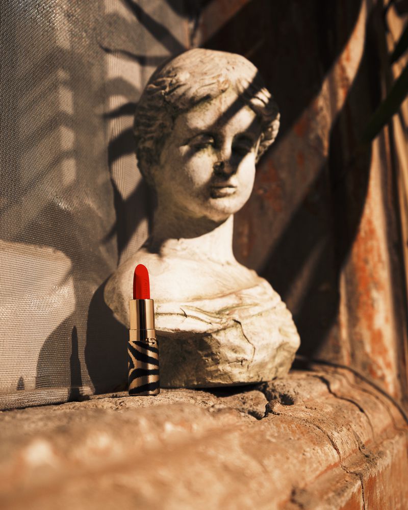 Close-up studio shot of red lipstick next to a stone bust on a textured surface, with shadows creating a sense of depth.