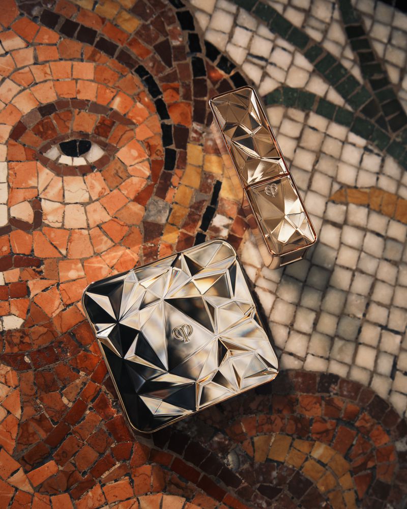Beauty still life featuring a faceted compact and lipstick on a mosaic background. The composition highlights textures and geometric designs.
