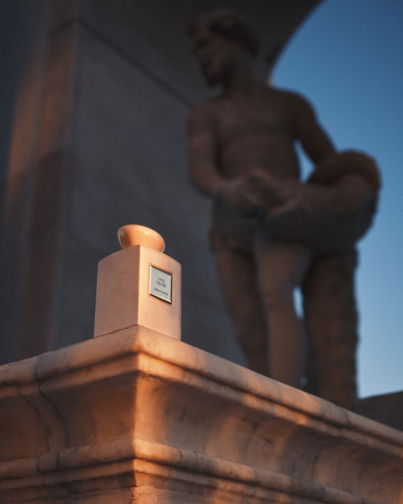 Elegant perfume bottle atop a stone pedestal, bathed in golden light, with a blurry statue in the background against a blue sky.