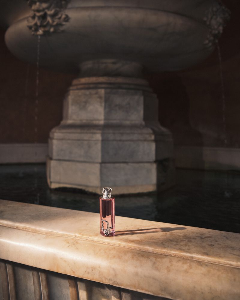 Perfume bottle with pink liquid sits on marble ledge near fountain. Perfume bottle with elegant silver cap and ornate logo.