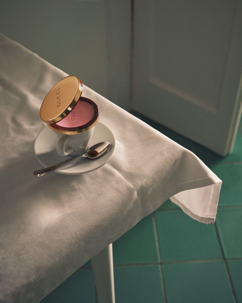 Close-up of gold Gucci powder compact on a white espresso cup saucer with a spoon, set on a table covered with a white tablecloth.