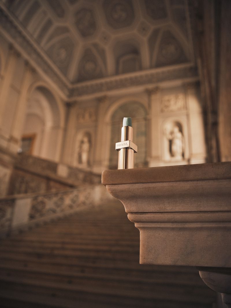 Prada lipstick tube on a marble ledge in a grand hall with a vaulted ceiling and staircase, product shot, soft lighting, neutral color palette.