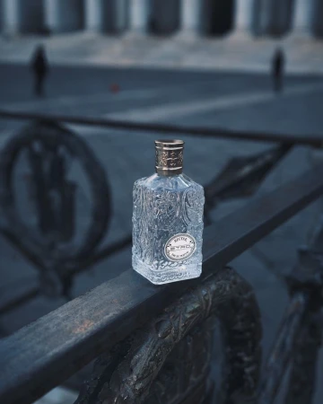 A clear glass perfume bottle with gold accents sits on an ornate black metal railing, with blurry buildings in the background.