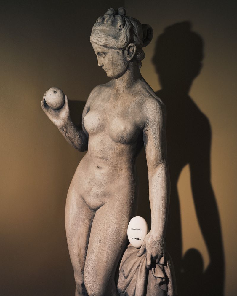 Classical statue holds an apple and a Chanel soap bar.  Stone sculpture of a female figure against a plain wall. Dark, moody lighting.