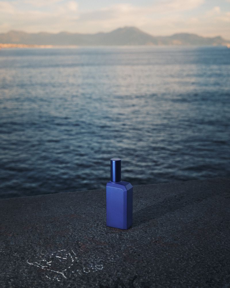 A blue perfume bottle sits on a concrete surface with a blurred ocean and mountain background under an overcast sky.