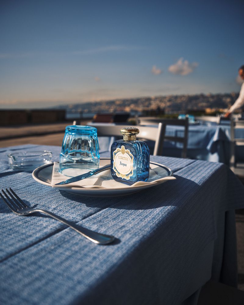 Elevated view of a perfume bottle next to a glass and silverware on a blue table cloth with a scenic background of the city.
