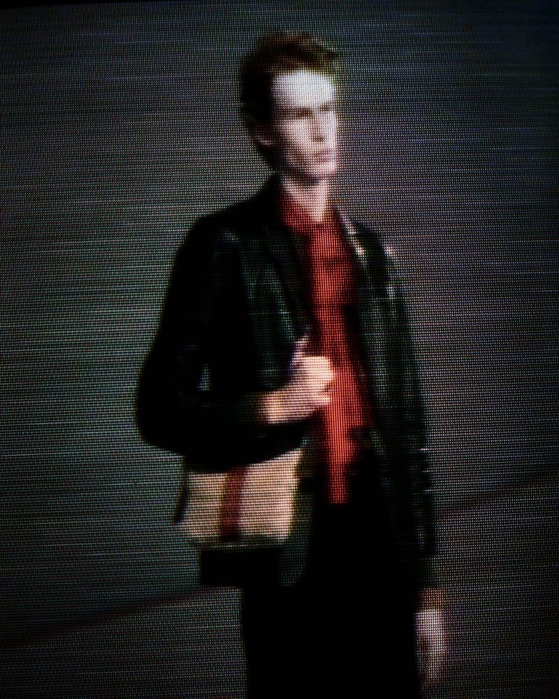 Man in black blazer and red shirt, carrying bag, stands in front of a neutral background in an editorial fashion shot.
