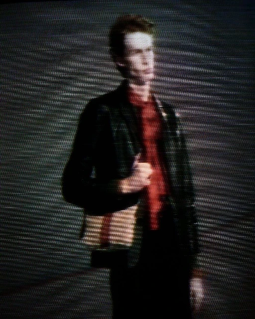 Man in black blazer and red shirt, carrying bag, stands in front of a neutral background in an editorial fashion shot.
