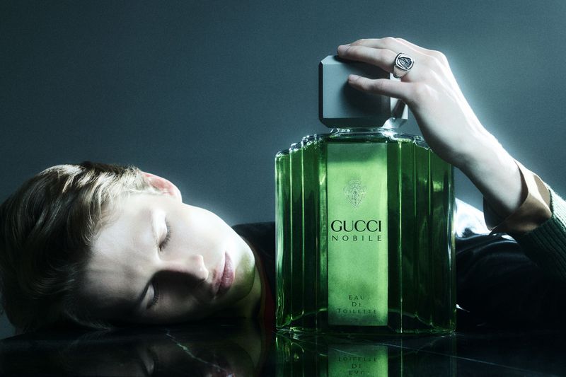 A green glass Gucci Noblie Eau de Toilette perfume bottle rests on a reflective surface with a male model lying beside it.