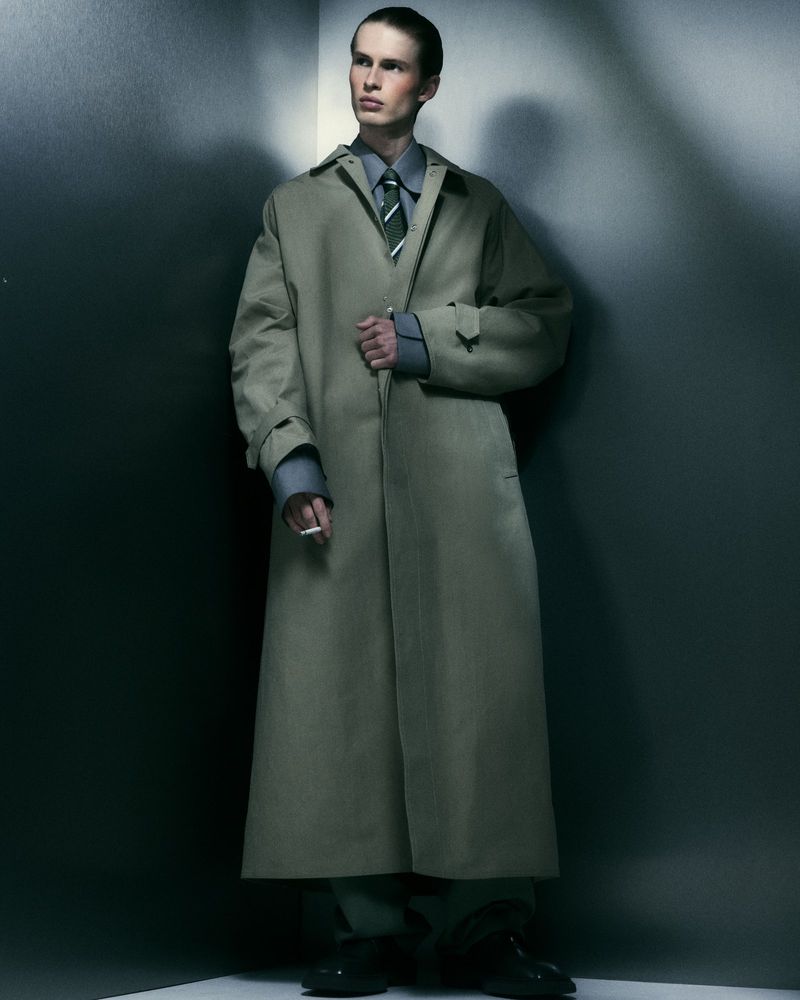 An adult male stands in a photo studio wearing a trench coat, shirt, tie, and trousers, holding a cigarette in his left hand. The backdrop is a neutral grey tone.