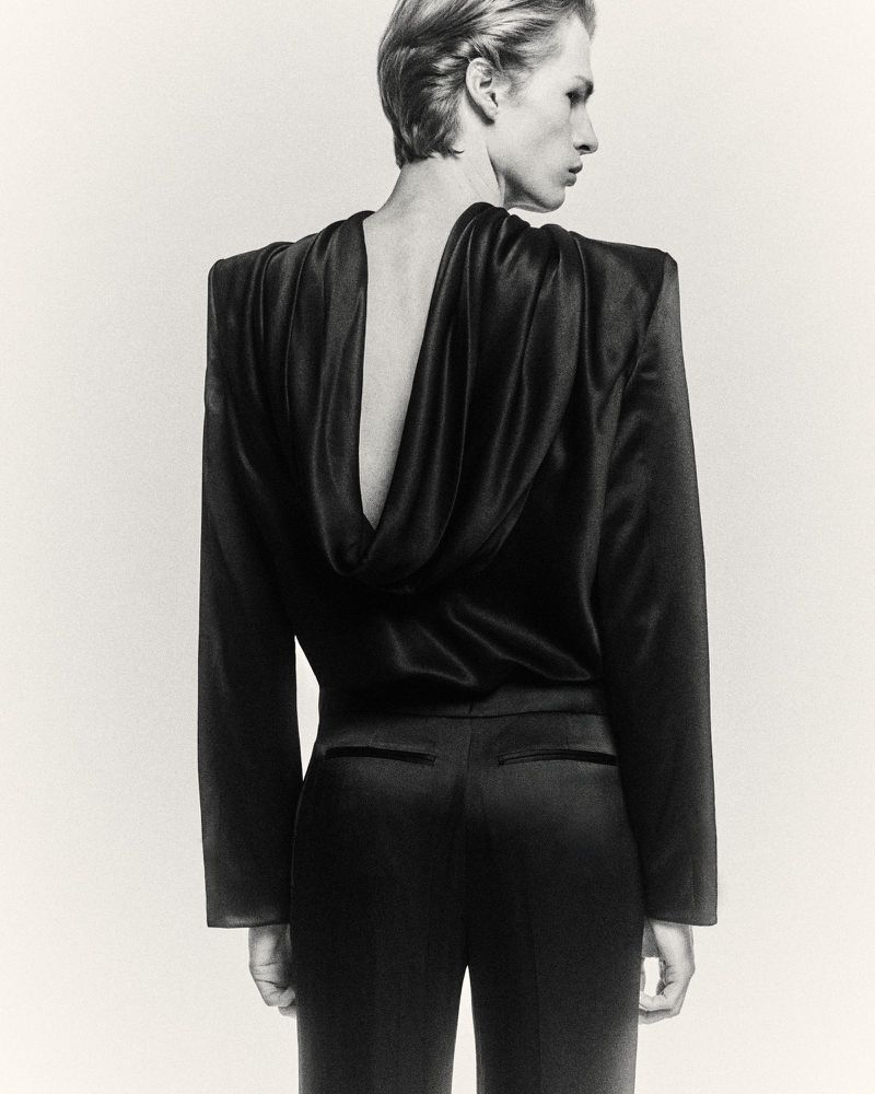 Monochrome fashion shot of a slender adult model in a dark draped top and trousers, showcasing a sleek backless design and elegant silhouette.