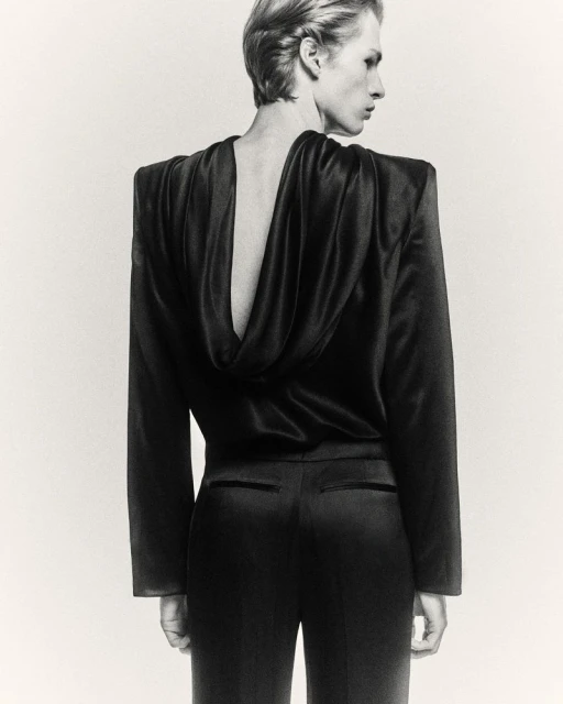 Monochrome fashion shot of a slender adult model in a dark draped top and trousers, showcasing a sleek backless design and elegant silhouette.
