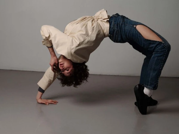 Person in a bridge pose wearing ripped jeans and a beige shirt against a grey background in a brightly lit indoor environment.