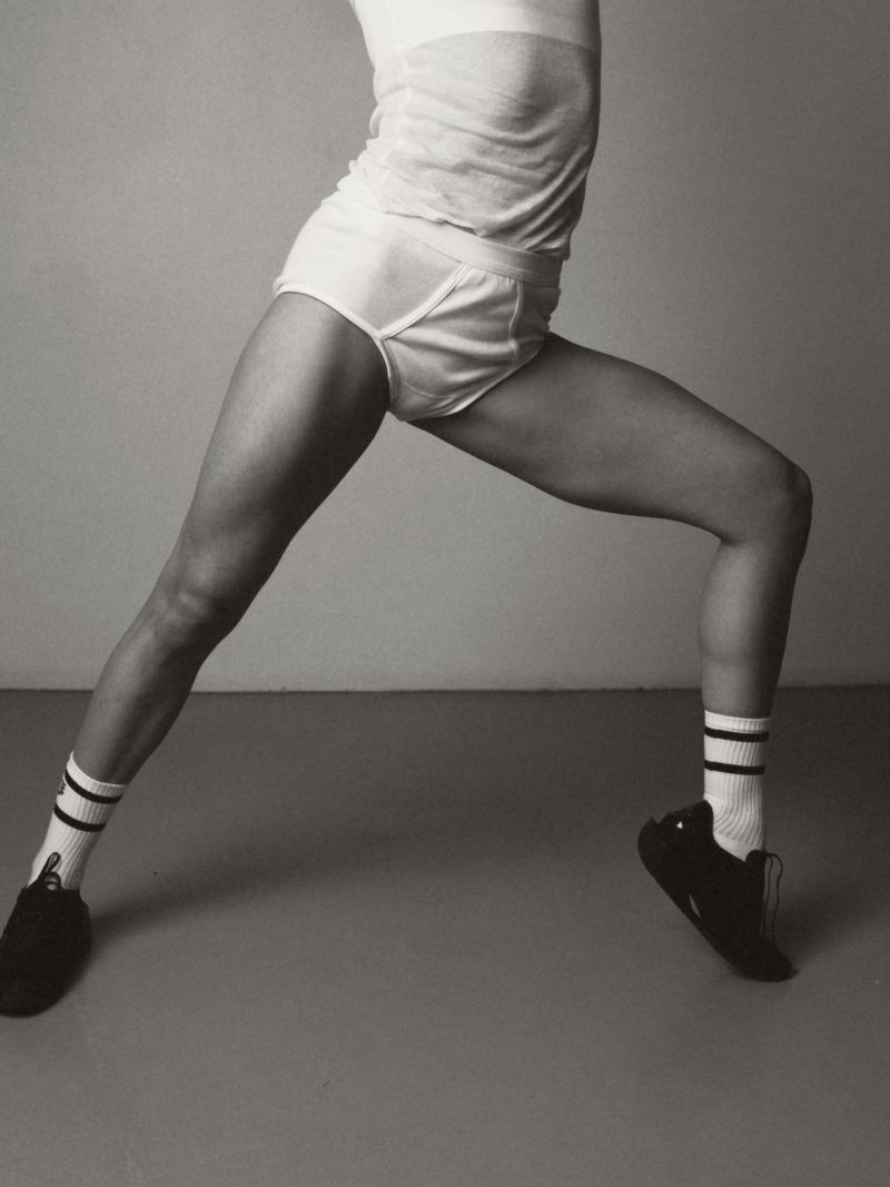 Monochromatic studio shot of an athletic woman in sportswear doing a yoga pose, wearing socks and athletic shoes against a grey background.