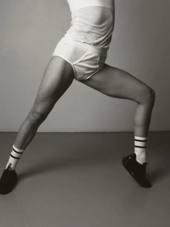 Monochromatic studio shot of an athletic woman in sportswear doing a yoga pose, wearing socks and athletic shoes against a grey background.