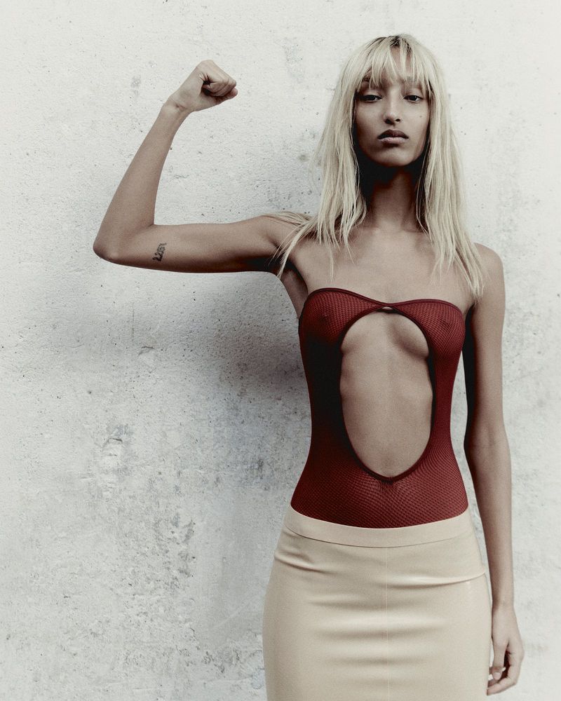 Woman in red bodysuit and tan skirt poses with raised arm against textured wall in a studio fashion shot. Portrait orientation.