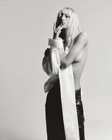 Black-and-white studio portrait of a topless adult female with blonde hair posing with an oversized white shirt and black pants.