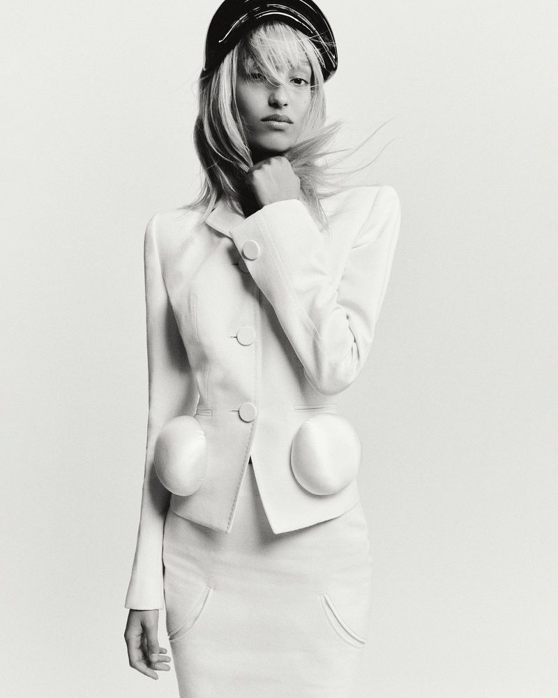 Monochrome fashion shot of woman in white skirt suit and black cap. Model's hand near face with neutral expression, light background.