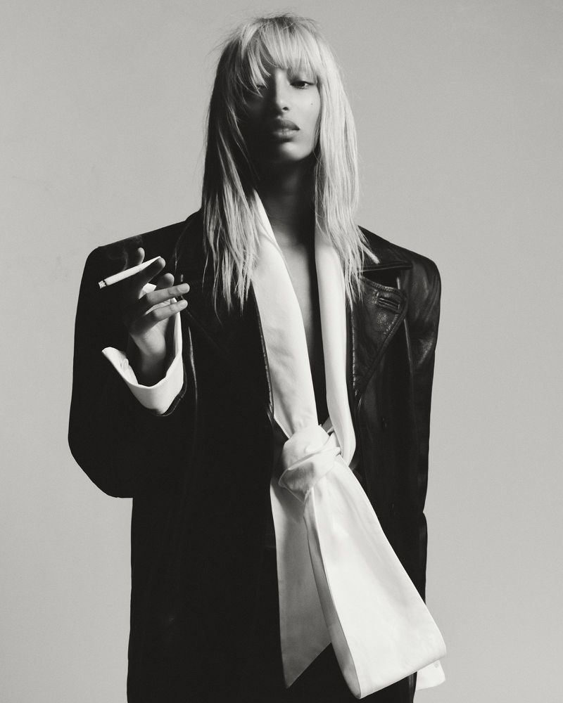 Monochrome portrait of a slender adult female with blonde hair, wearing a black jacket and white sash, holding a cigarette indoors.