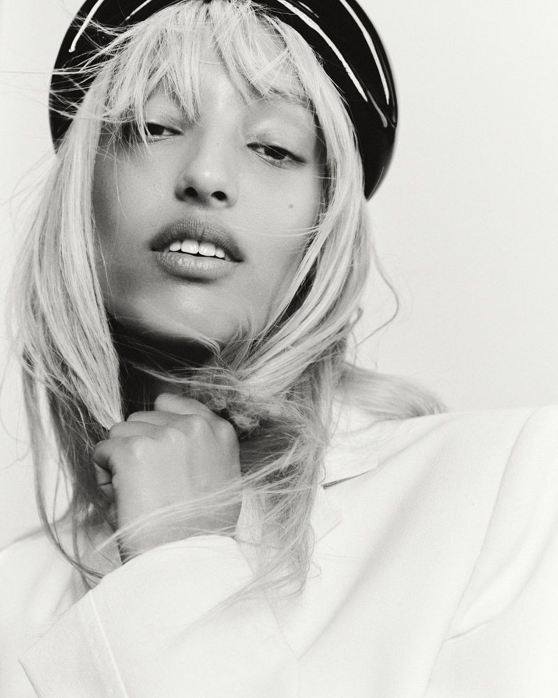 Close-up portrait of a young adult female model with blonde hair and a black headband, wearing a white jacket against a white background.