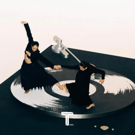 Two barefoot models strike expressive dance poses on a black and white vinyl record, with a turntable arm, against a white backdrop.