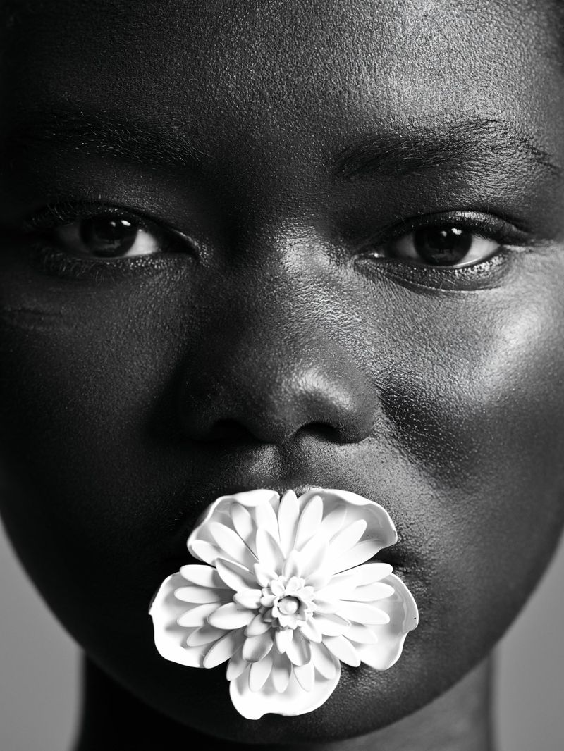 Close-up portrait of an African American woman with dark skin, a flower in her mouth, captured in black and white with a soft gray background.